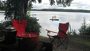 Camp kitchen, folding camping chairs, kayaks on lake -...